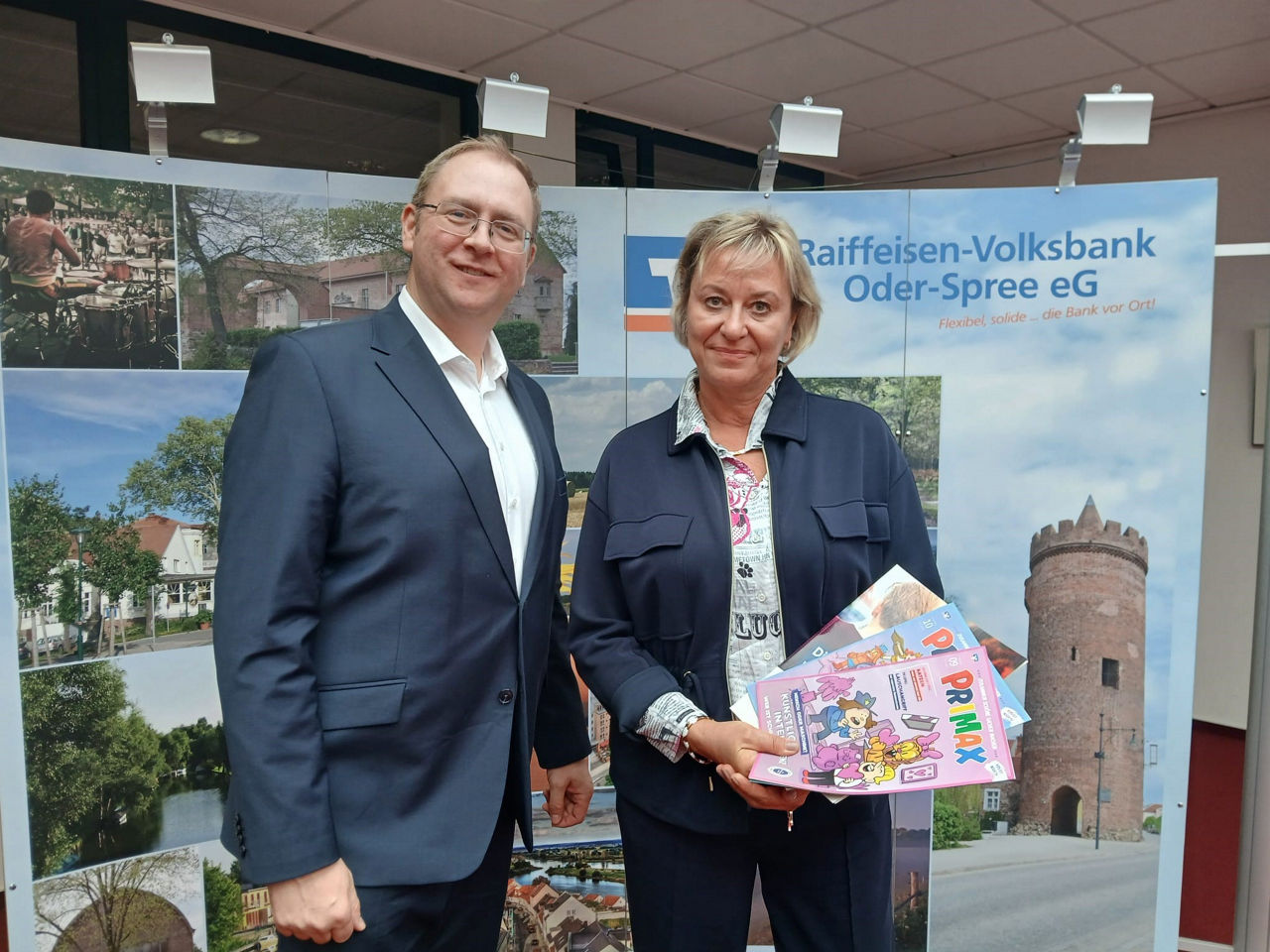 Mann und Frau halten mehrere PRIMAX-Magazine vor dem Aufsteller der Raiffeisen-Volksbank Oder-Spree eG.
