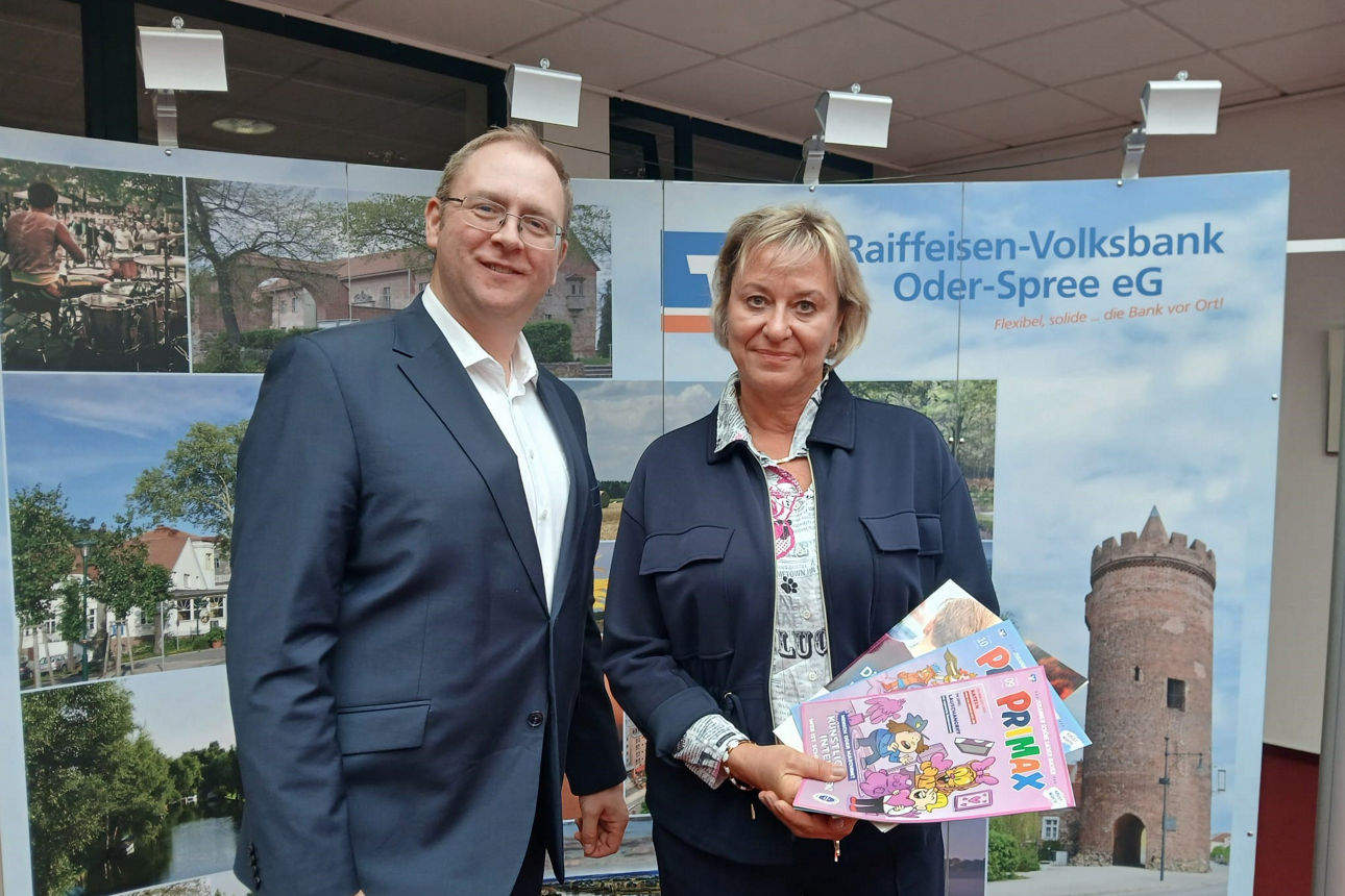 Mann und Frau halten mehrere PRIMAX-Magazine vor dem Aufsteller der Raiffeisen-Volksbank Oder-Spree eG