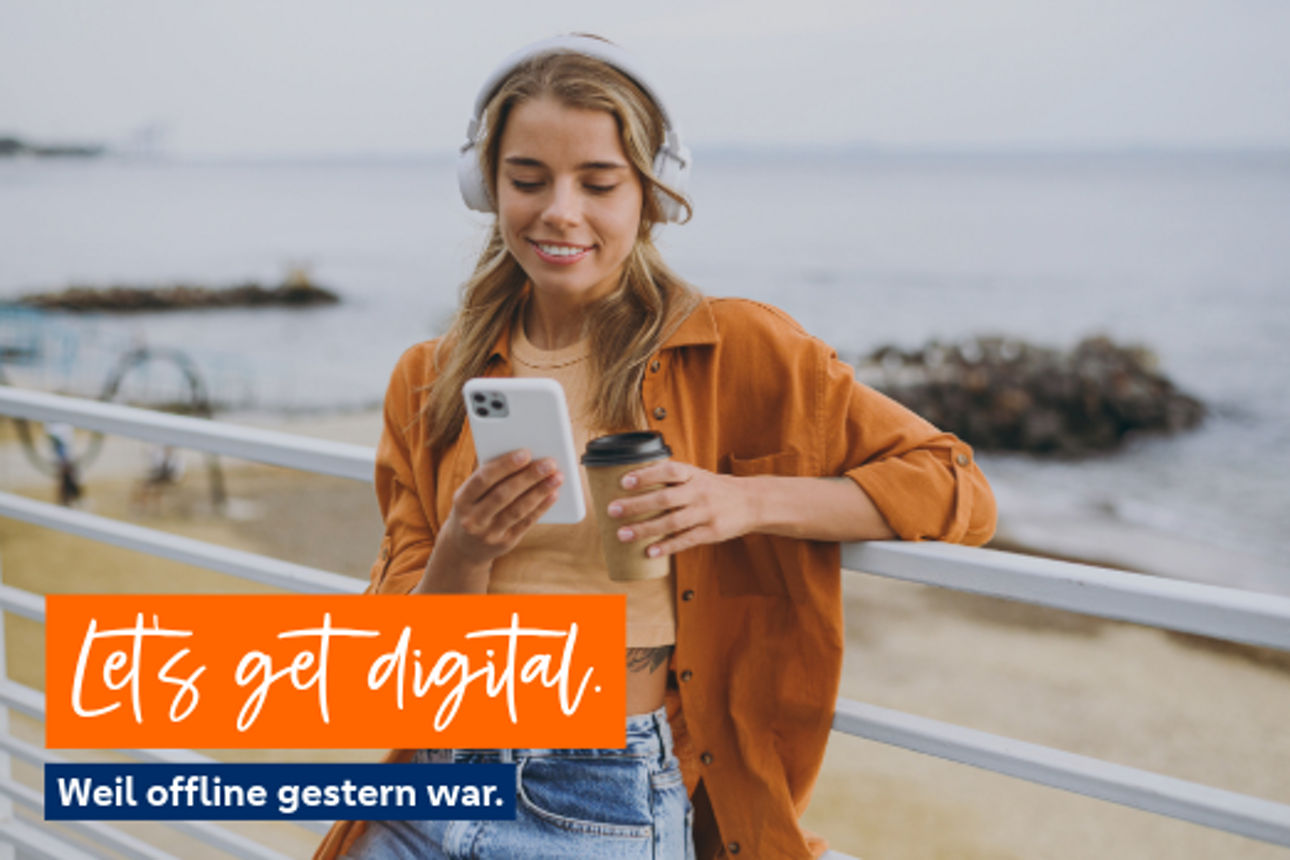 Eine Frau mit Kopfhörern am Strand mit Handy und Kaffeebecher in der Hand