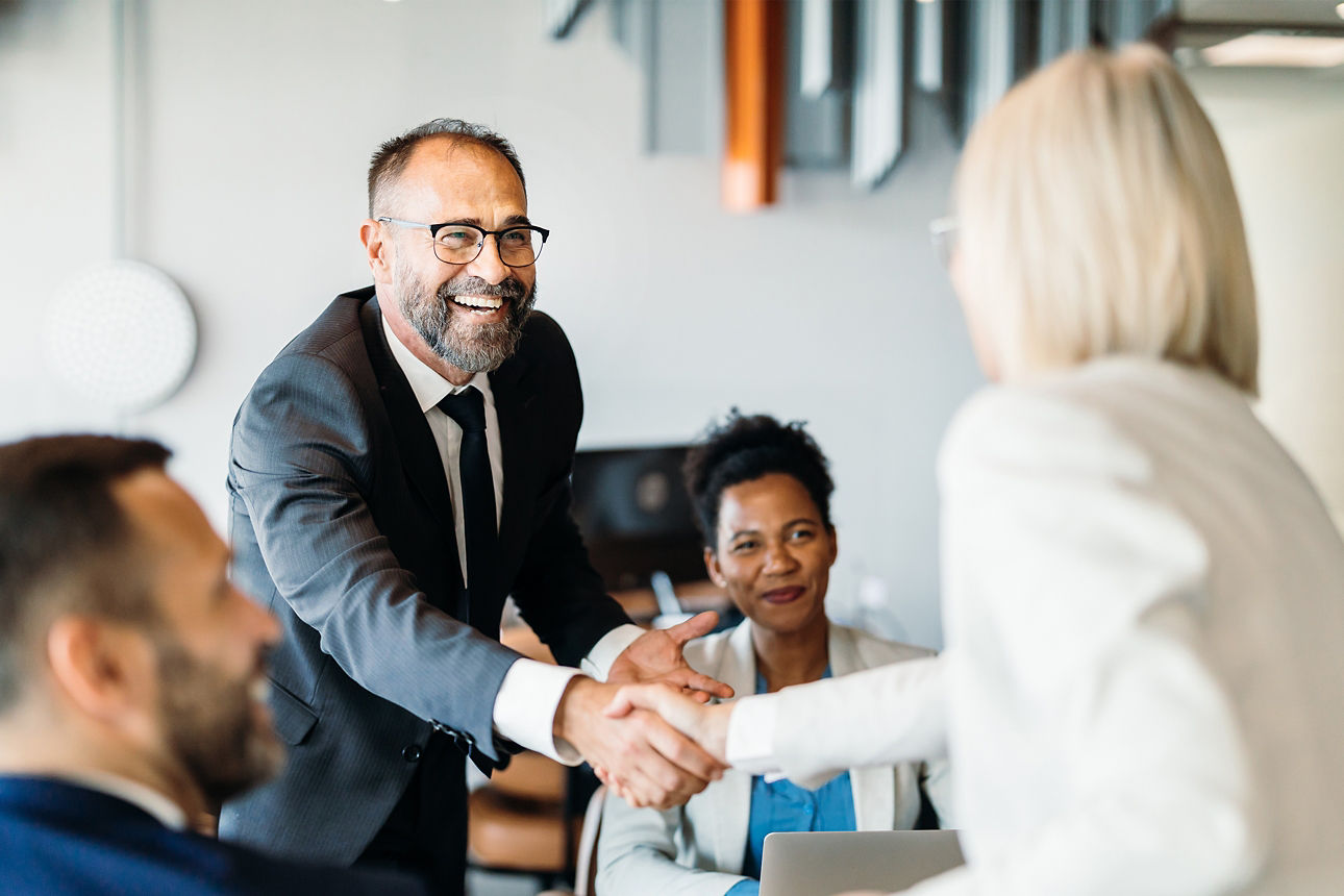 Meeting-Situation.Ein Mann schüttelt einer Frau freundlich lachend die Hand. Zwei weitere Personen sitzend lächelnd am Tisch.