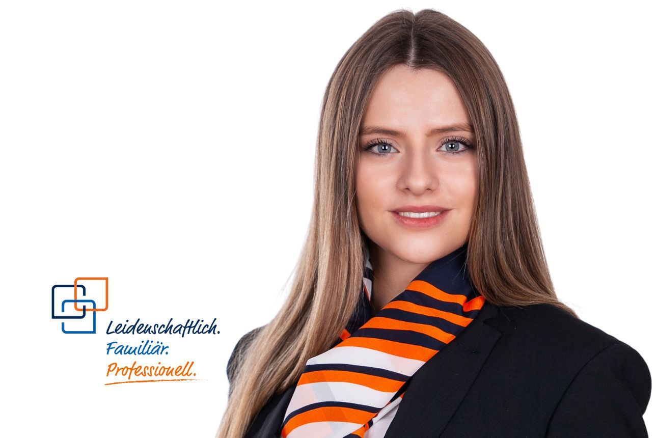 Linda Semtner, eine junge Frau mit langen braunen Haaren und blau-grauen Augen, lächelt freundlich und trägt einen dunklen Blazer mit einem gestreiften orange-blau-weißen Schal.