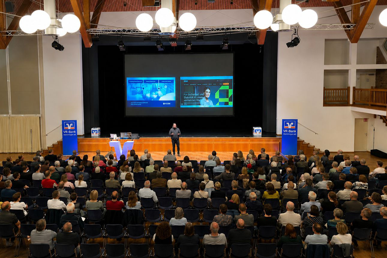 Vortragende steht auf der Bühne vor großem Publikum im Saal mit zwei Projektionsflächen und Bannern der VR-Bank Memmingen