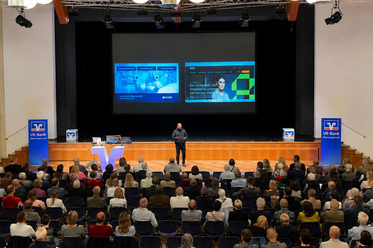 Vortragende steht auf der Bühne vor großem Publikum im Saal mit zwei Projektionsflächen und Bannern der VR-Bank Memmingen