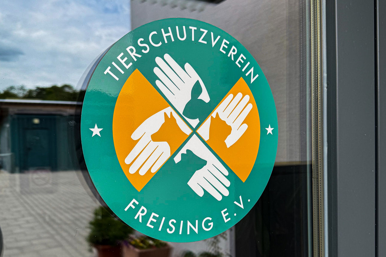 rundes Logo klebt auf Eingangstür; auf grün-orangenem Hintergrund sind vier weiße Hände abgebildet, die sich schützend über Silhouetten von Tierköpfen halten