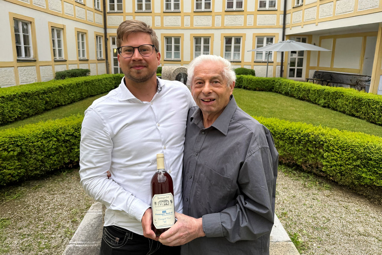 EIn junger und ein alter Mann halten sich vertraut im Arm; im Vordergrund halten sie gemeinsam eine Flache Wein in die Kamera