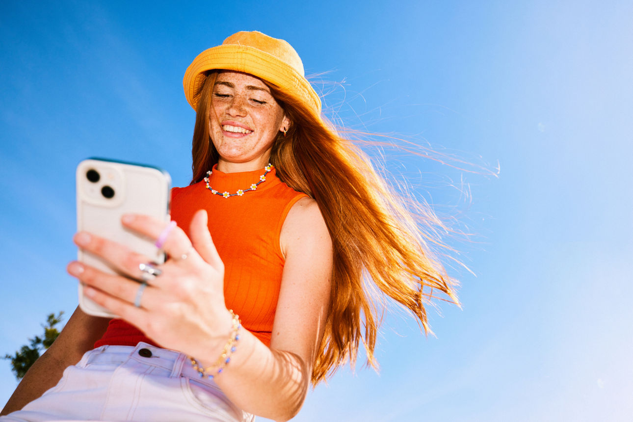 Eine junge Frau mit orangem Oberteil und gelbem Hut lächelt, während sie bei Sonnenschein auf ihr Smartphone schaut.