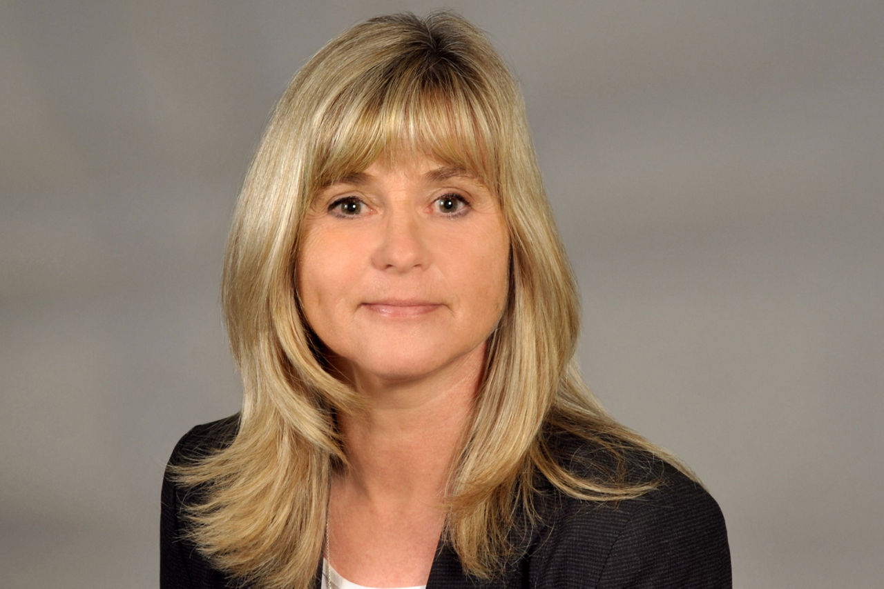 Frau mit schulterlangem blondem Haar, trägt schwarzen Blazer und weiße Bluse, vor grauem Hintergrund