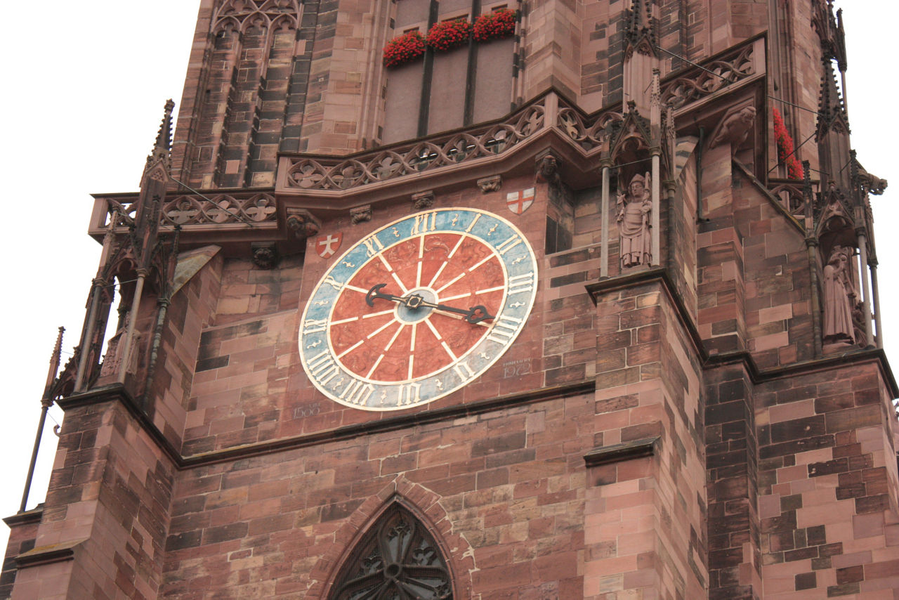 Eine große runde Uhr mit römischen Ziffern ist an einem roten, gotischen Kirchturm befestigt.