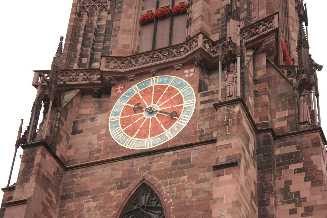 Eine große runde Uhr mit römischen Ziffern ist an einem roten, gotischen Kirchturm befestigt.