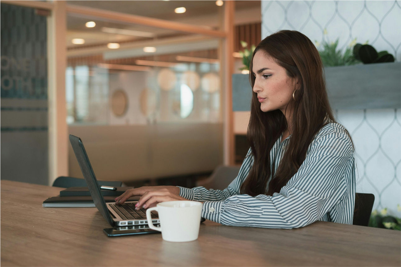Eine Frau sitzt an einem Tisch, vor ihr ein Laptop und eine Tasse. 