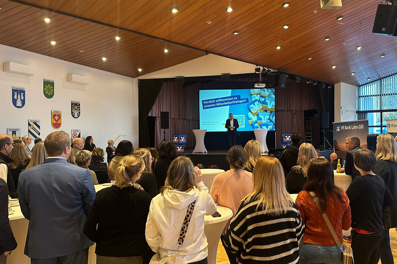 Menschen stehen in einem Saal und hören einem Redner auf einer Bühne mit Präsentation zu, an der Wand sind mehrere Wappen angebracht.