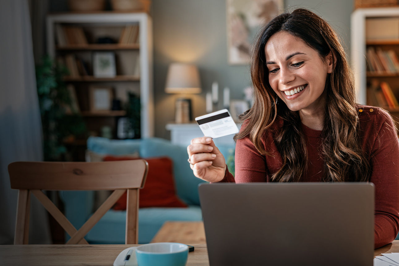 Eine junge lächelnde Frau sitzt vor Ihrem Laptop und hält eine Kreditkarte in der Hand