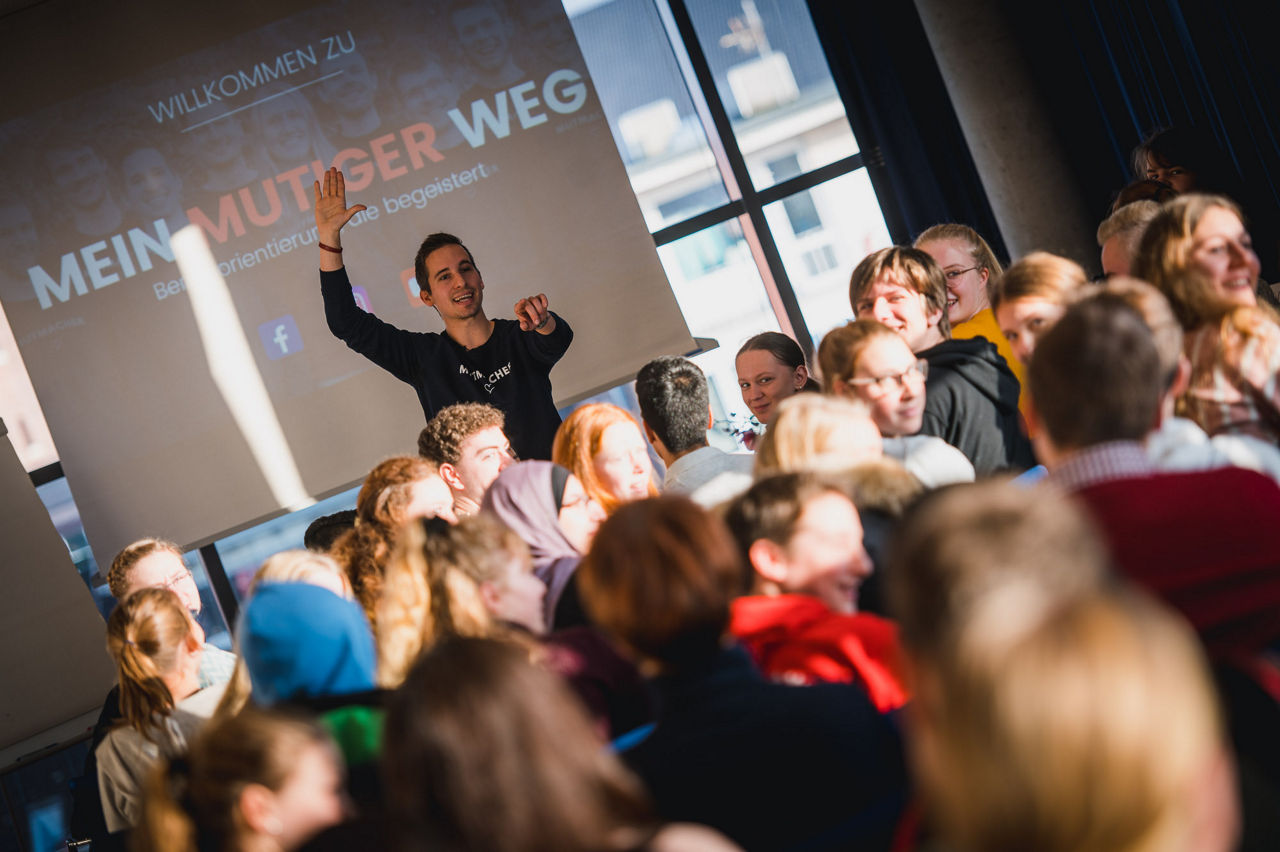 Ein Mann steht vor einer Schulklasse. Im Hintergrund ist der Banner von "Mein mutiger Weg" zu erkennen.