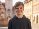 KI-generiertes Porträt eines lächelnden Jugendlichen mit kurzen braunen Haaren und dunklem Hoodie. Die Person blickt direkt in die Kamera. Im unscharfen Hintergrund sind historische Gebäude und eine Altstadtstraße bei warmem Tageslicht zu sehen.