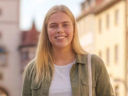 KI-generiertes Porträt einer lächelnden jungen Frau mit langen blonden Haaren. Sie trägt ein helles Oberteil, eine olivgrüne Jacke, eine Halskette und eine Schultertasche. Sie blickt in die Kamera. Im unscharfen Hintergrund sind historische Gebäude und eine Altstadtstraße bei warmem Tageslicht zu sehen.