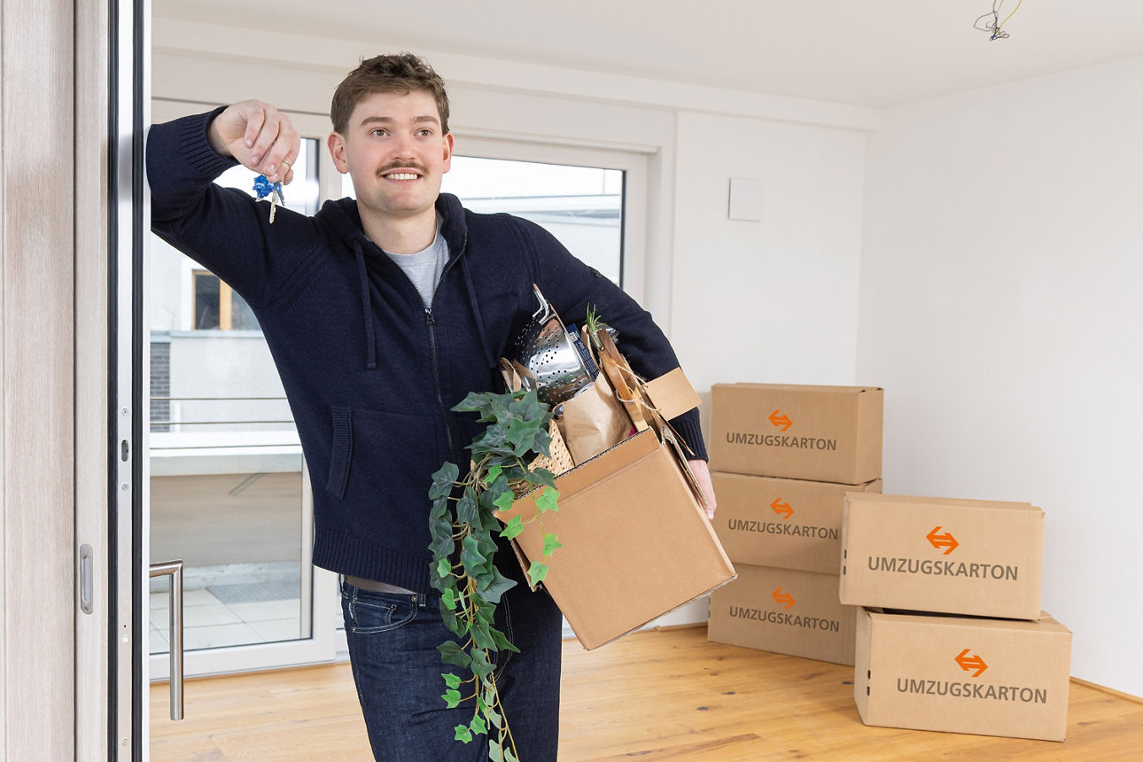 Junger Mann steht mit Schlüssel und Umzugskartons in seiner ersten Wohnung – Meine Junge Volksbank
