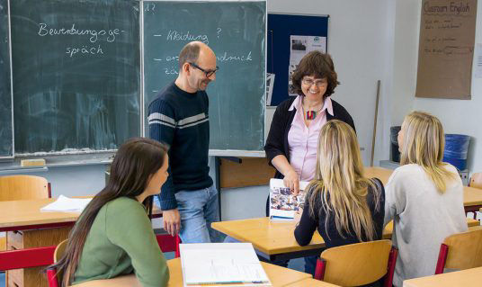 Das Bild zeigt eine Situation in einer Schulklasse eine Lehrerin und ein Lehrer stehen vor drei Schülerinnen.