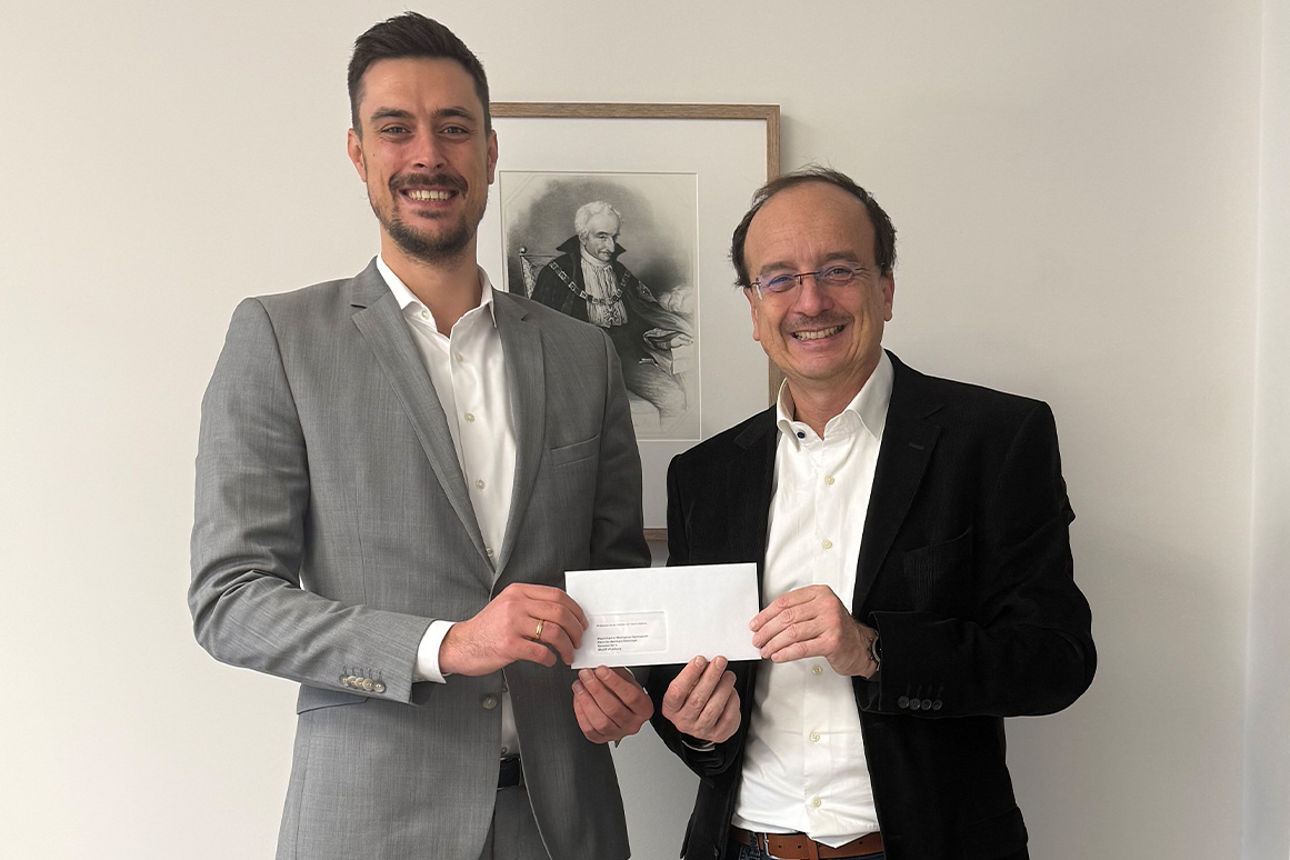 Zwei Männder lächeln in die Kamera. Sie halten einen Briefumschlag in der Hand.