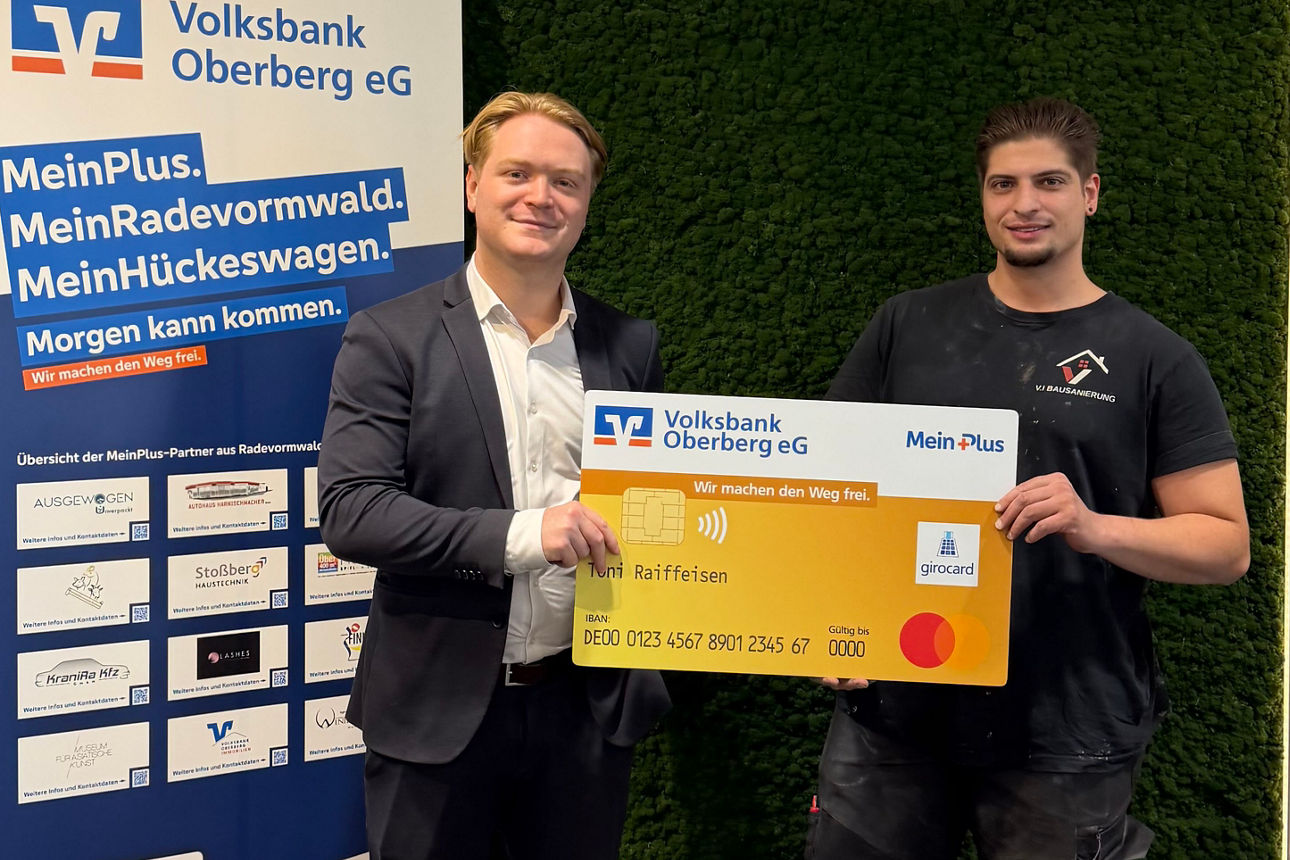 Zwei Männer lächeln in die Kamera und halten gemeinsam eine übergroße, symbolische Bankkarte der Volksbank Oberberg.