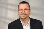 Christoph Machmerth hat kurze dunkle Haare, einen Dreitagebart und eine eckige Brille. Er trägt einen Anzug.