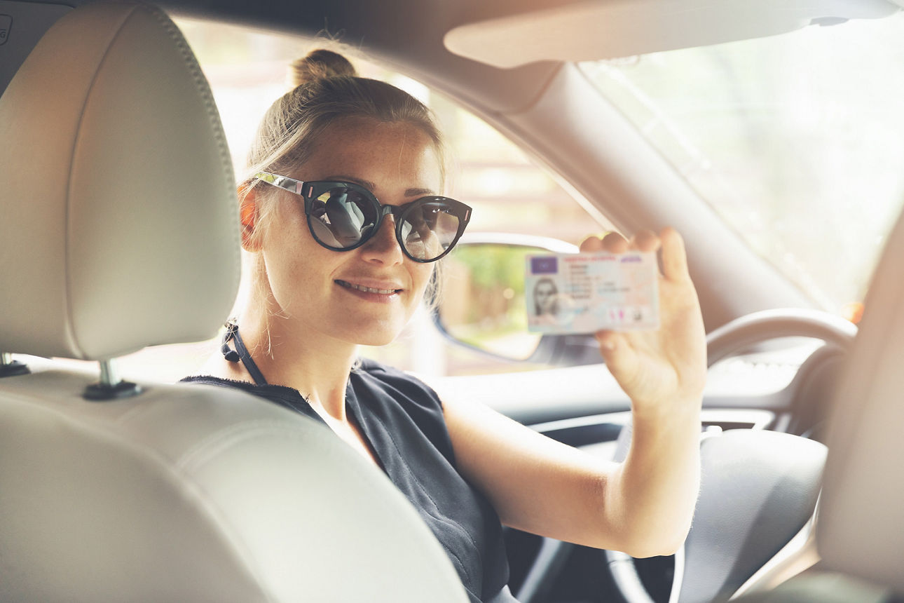Mädchen sitzt lächelnd im Auto und zeigt ihren Führerschein