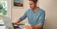 Ein Mann sitzt am Laptop, in der linken Hand hält er ein Handy 