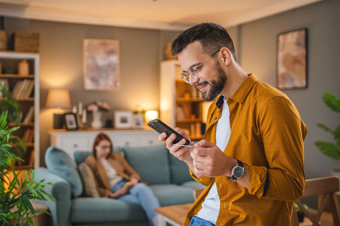 Mann steht in Wohnzimmer und blickt auf Handy und Bankkarte