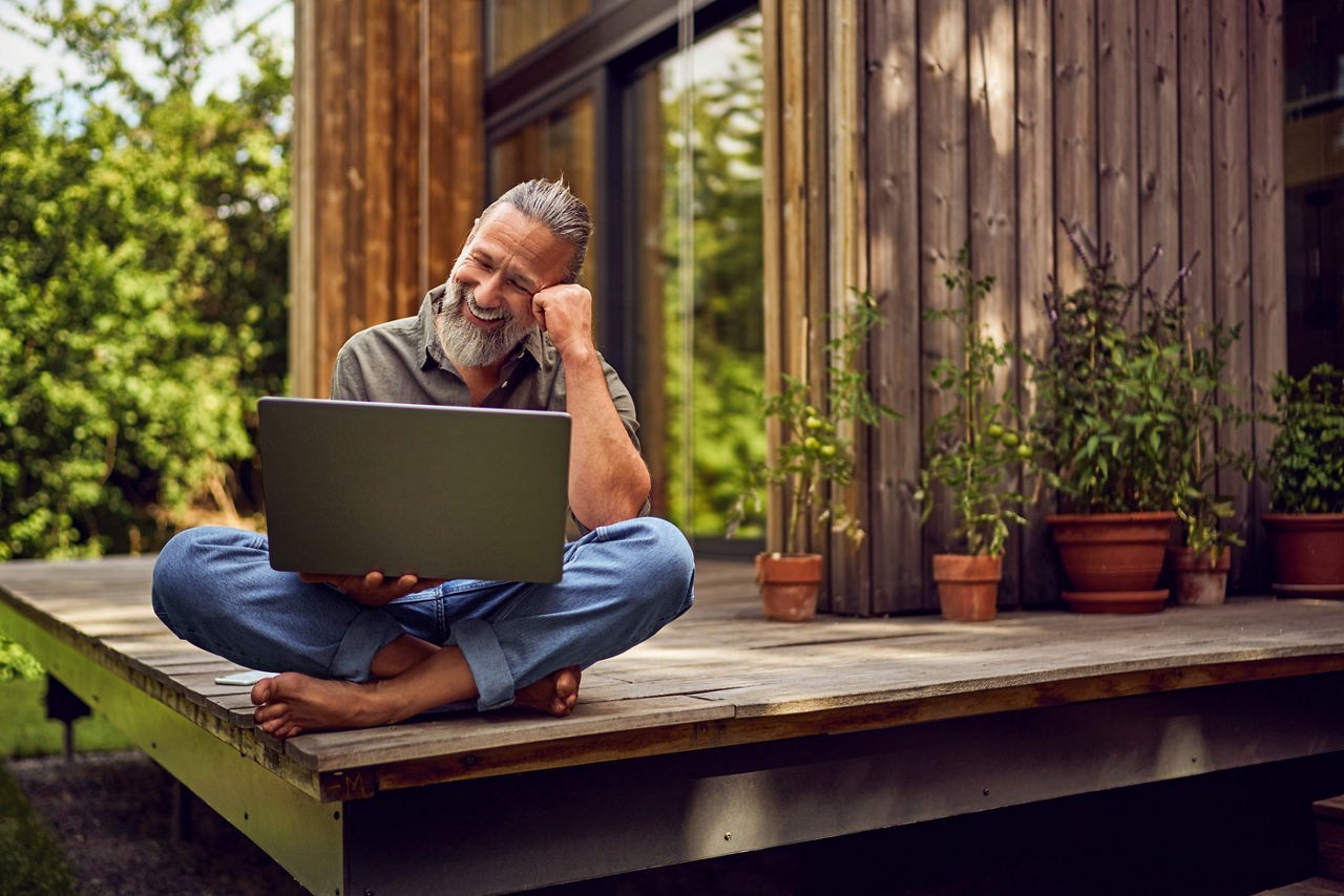 Lachender Mann sitzt mit seinem Laptop auf dem Schoß auf der Terrasse zu seinem Haus