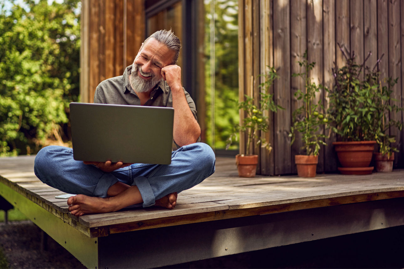 Lachender Mann sitzt mit seinem Laptop auf dem Schoß auf der Terrasse zu seinem Haus