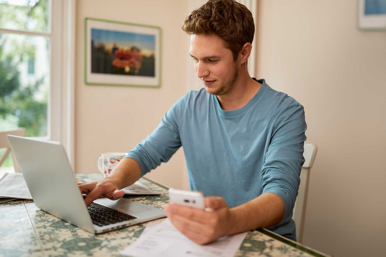 Junger Mann sitzt vor seinem Laptop mit seinem Handy in der Hand