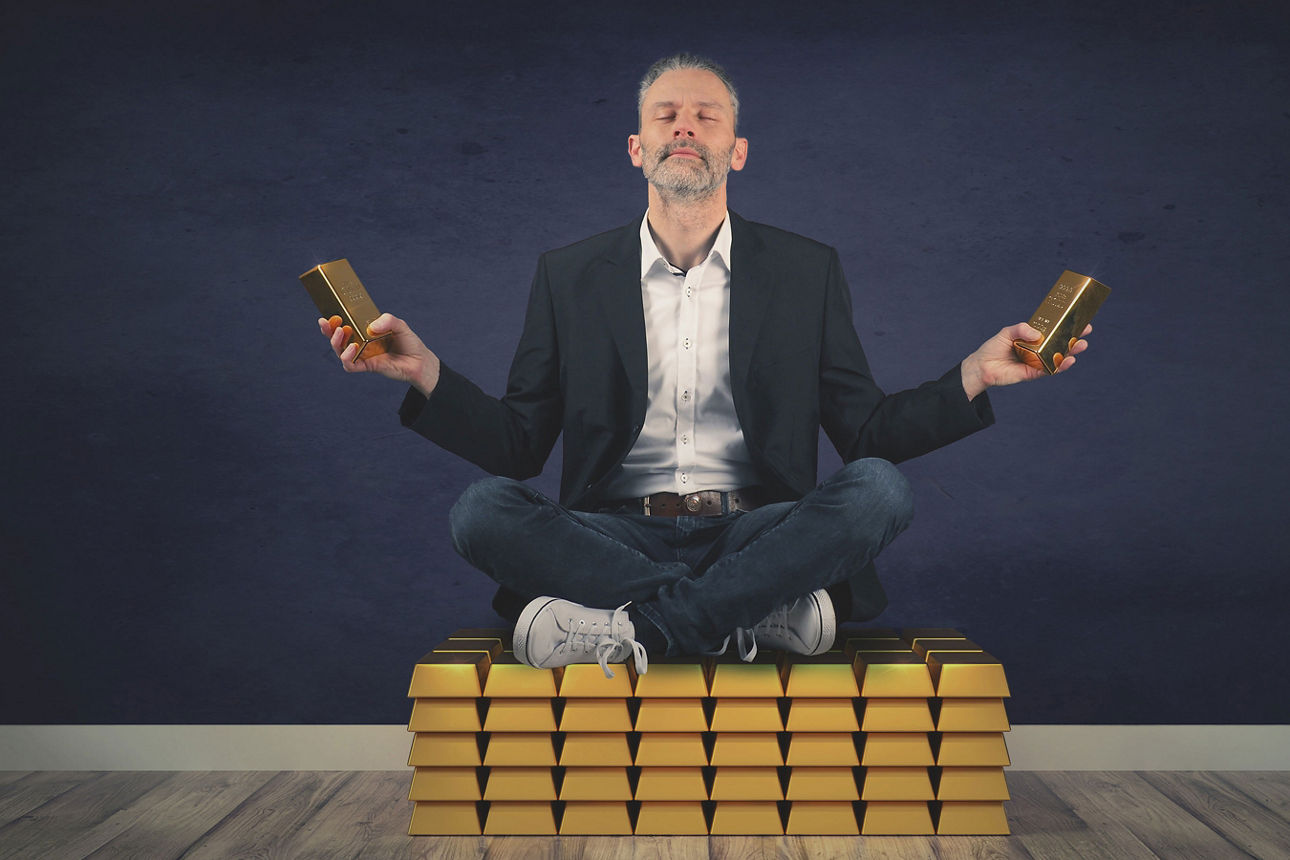 Mann sitzt im Schneidersitz auf gestapelten Goldbarren und hält in jeder Hand einen Goldbarren, Augen geschlossen.