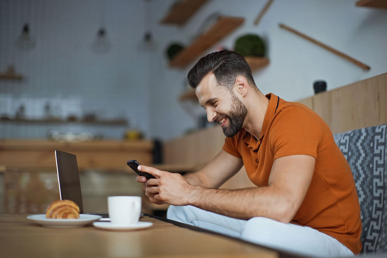 Mann sitzt entspannt am Tisch, schaut aufs Smartphone; Laptop, Kaffee und Croissant stehen daneben.