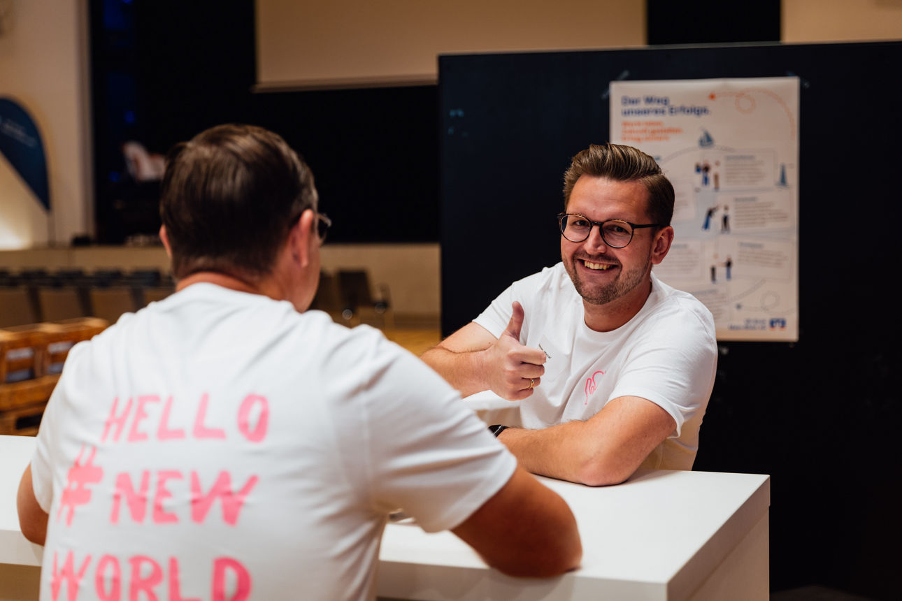 Ein Mann schaut lächelnd in die Kamera und hält den Daumen hoch. Man sieht auch, dass er mit einem anderen Mann an einem Tisch sitzt. Diesen sieht man nur von hinten.