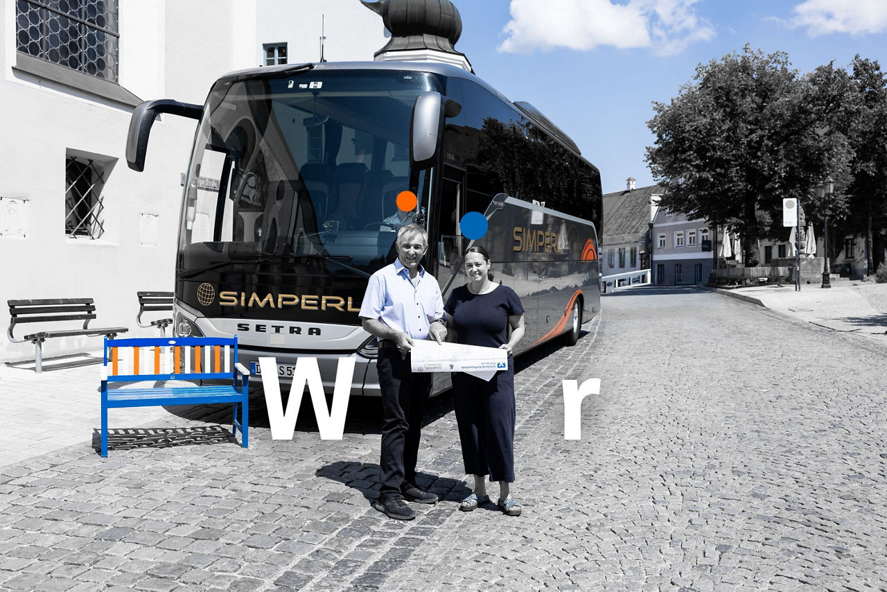 Ein Mann und eine Frau stehen vor einem Reisebus, links beim Bus steht eine Holzbank