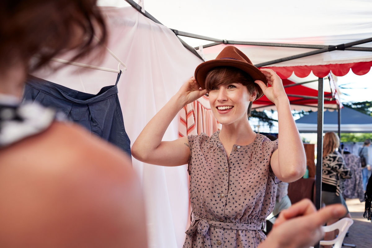 Cute woman trying on hat in front of girl friend, at small market