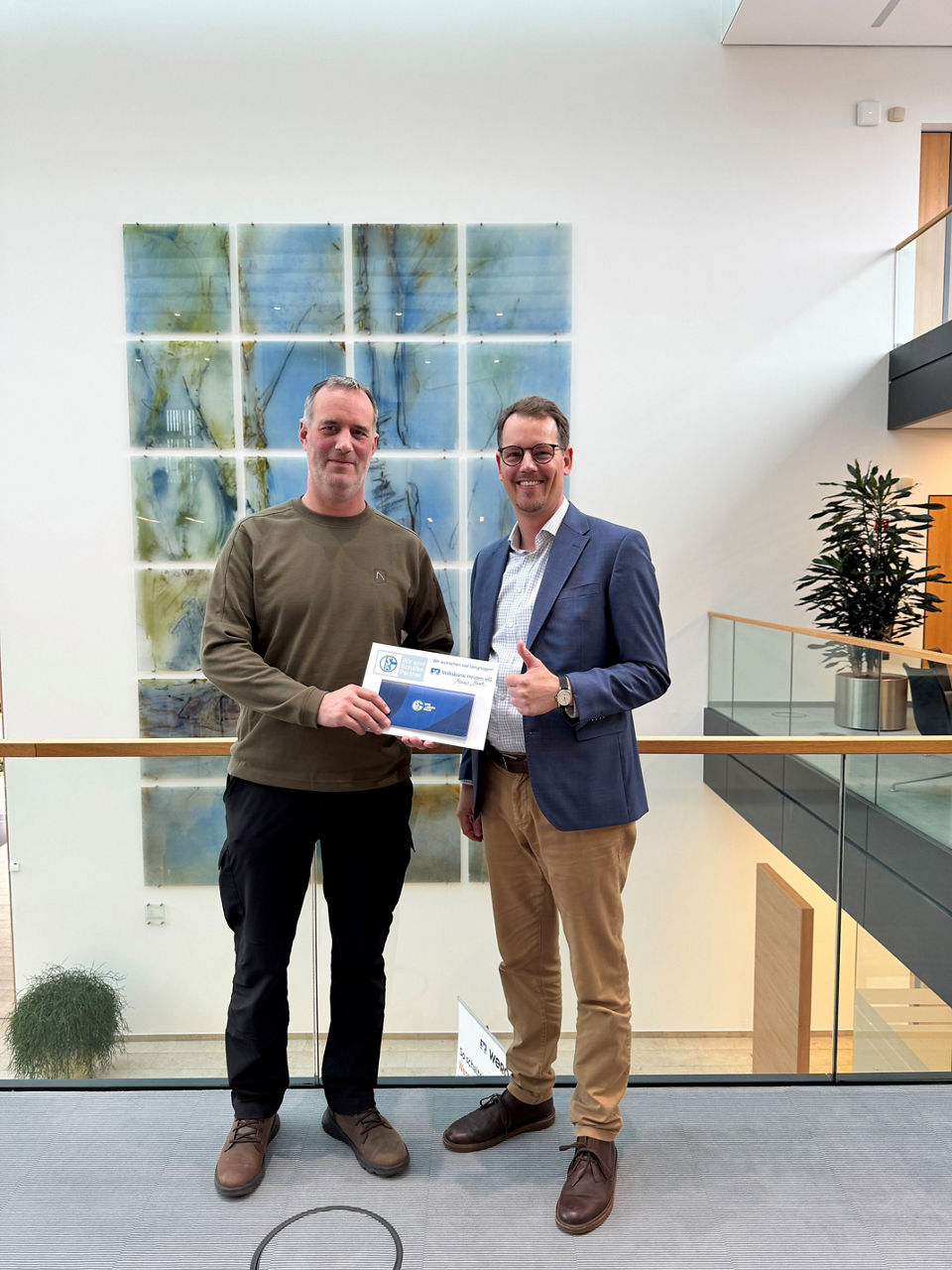 Zwei Personen lächeln in die Kamera, zeigen den Daumen hoch und halten gemeinsam ein Dokument in den Händen.