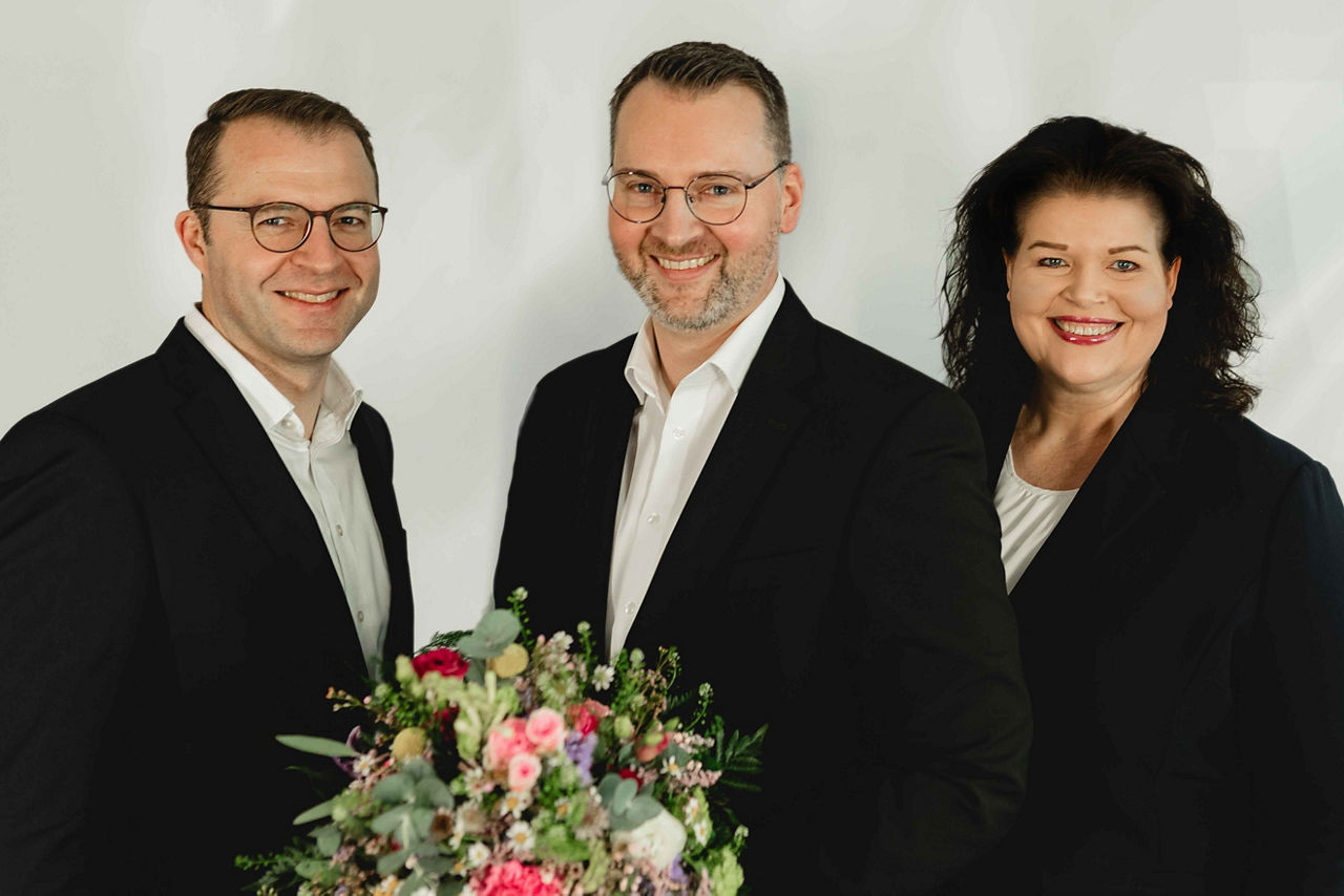 Zwei Vorstandsmitglieder und ein Mitarbeiter, freundlich lächelnd, mit Blumenstrauß – vor hellem Hinter