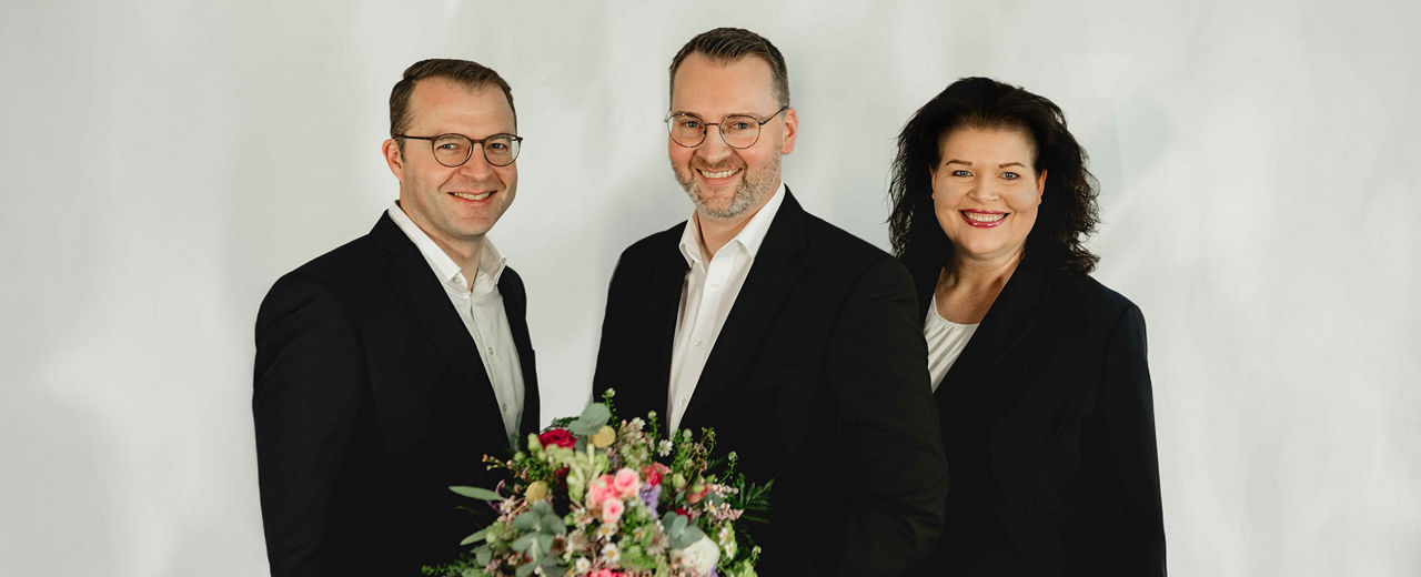 Zwei Vorstandsmitglieder und ein Mitarbeiter, freundlich lächelnd, mit Blumenstrauß – vor hellem Hinter