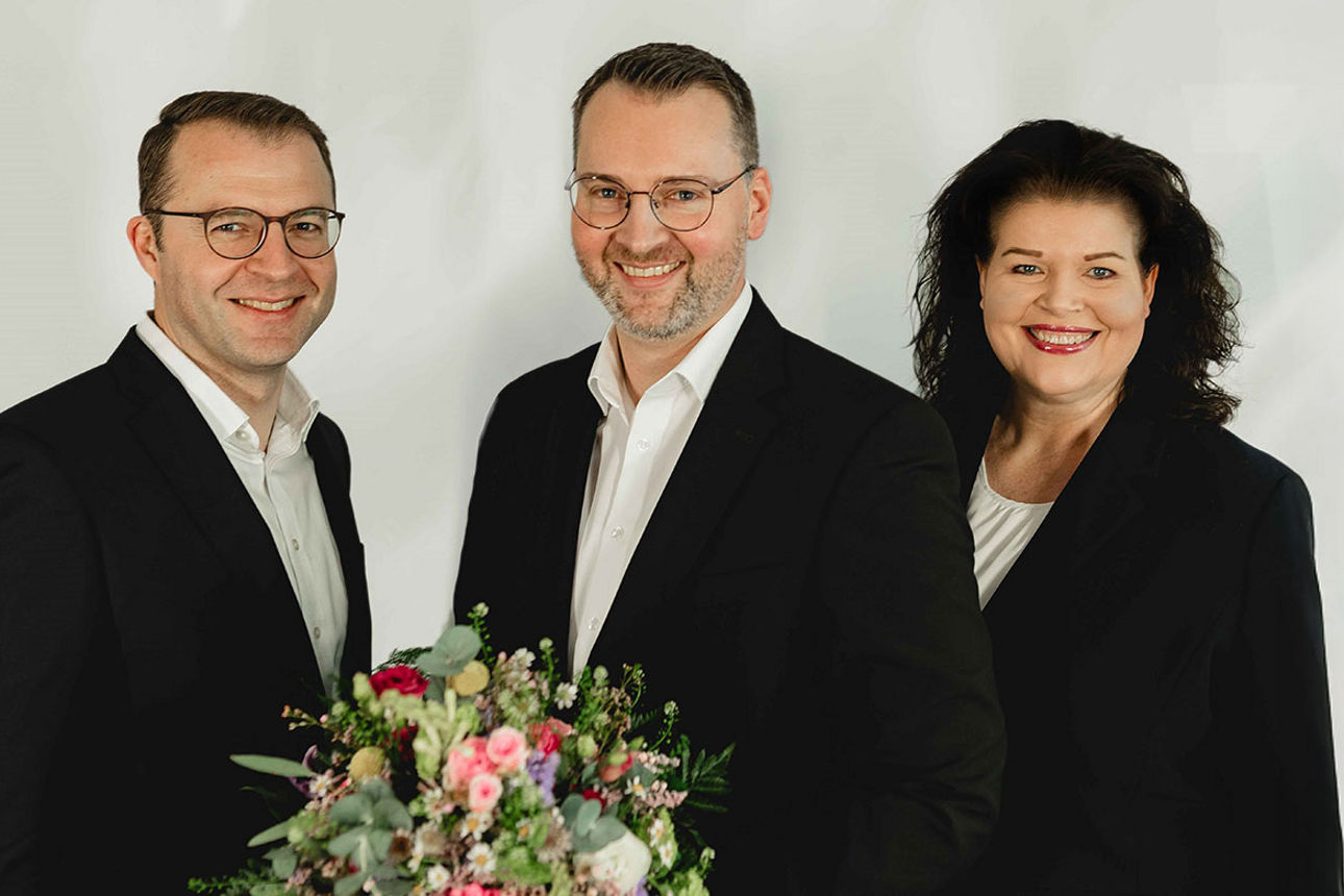 Zwei Vorstandsmitglieder und ein Mitarbeiter, freundlich lächelnd, mit Blumenstrauß – vor hellem Hintergrund