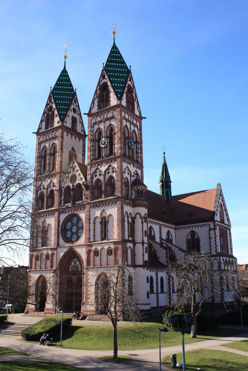 Das Bild zeigt eine Kirche mit zwei hohen Türmen, grün gedeckten Spitzdächern und einem großen runden Fenster über dem Hauptportal. Vor der Kirche befindet sich ein gepflegter Platz mit Wegen, Bäumen und Sitzbänken.