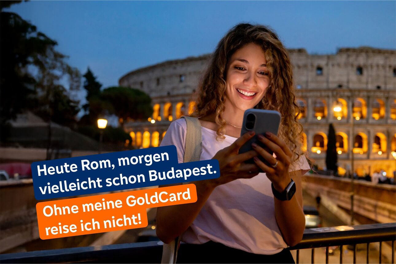 Junge Frau mit lockigem Haar steht abends vor dem beleuchteten Kolosseum in Rom und schaut auf ihr Smartphone.