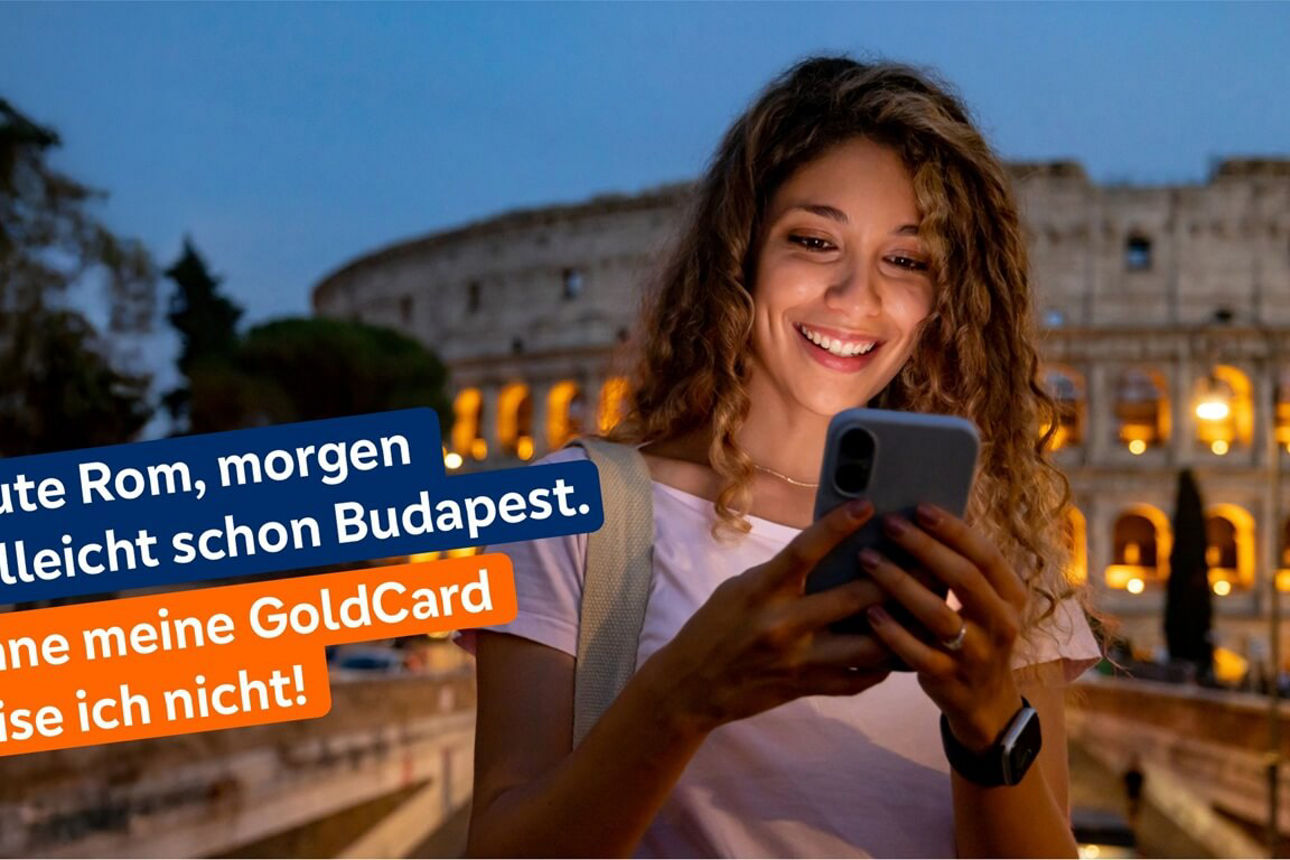 Junge Frau mit lockigem Haar steht abends vor dem beleuchteten Kolosseum in Rom und schaut auf ihr Smartphone.