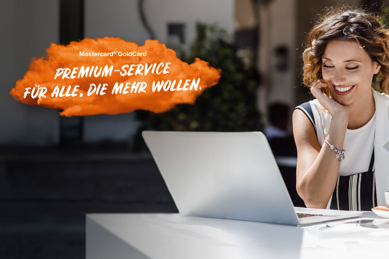 Lächelnde junge Frau sitzt mit ihrem Laptop am Tisch und rührt in ihrem Kaffee. Links im Bild steht auf einer orangenen Fläche "Mastercard® Gold: Premium Service für alle, die mehr wollen."