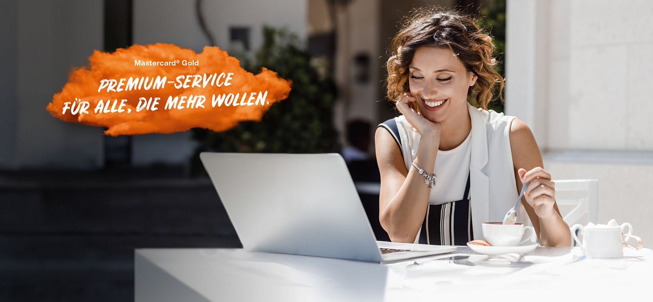 Lächelnde junge Frau sitzt mit ihrem Laptop am Tisch und rührt in ihrem Kaffee. Links im Bild steht auf einer orangenen Fläche "Mastercard® Gold: Premium Service für alle, die mehr wollen."