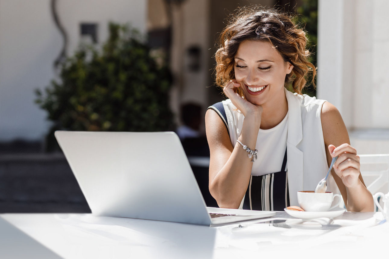 Lächelnde junge Frau sitzt mit ihrem Laptop am Tisch und rührt in ihrem Kaffee.