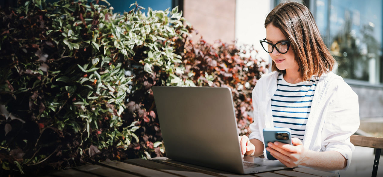 Junge Frau sitzt außen an einem Tisch mit ihrem Smartphone und ihrem Laptop..