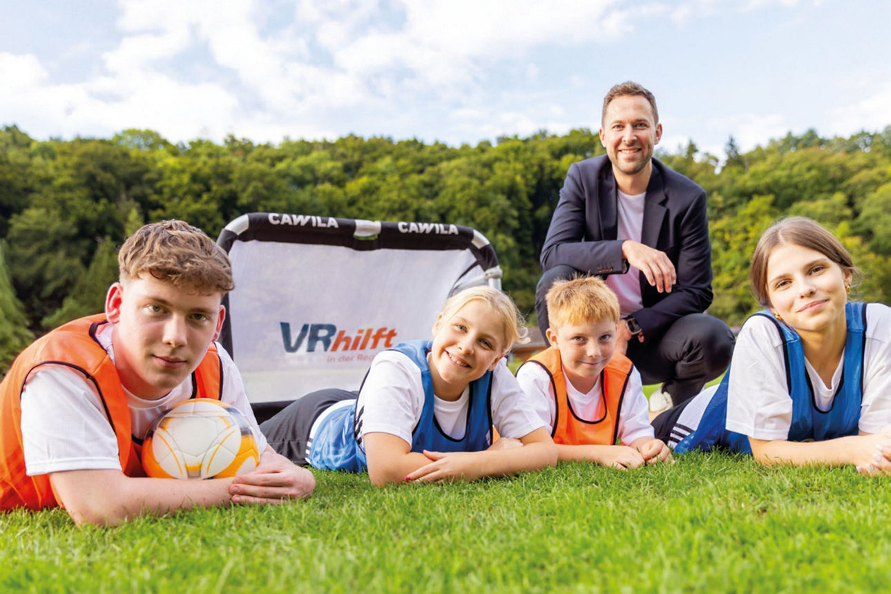 Kinder liegen lachend auf dem Rasen vor einem Fußballtor, während ein Trainer hinter ihnen stolz in die Kamera schaut.
