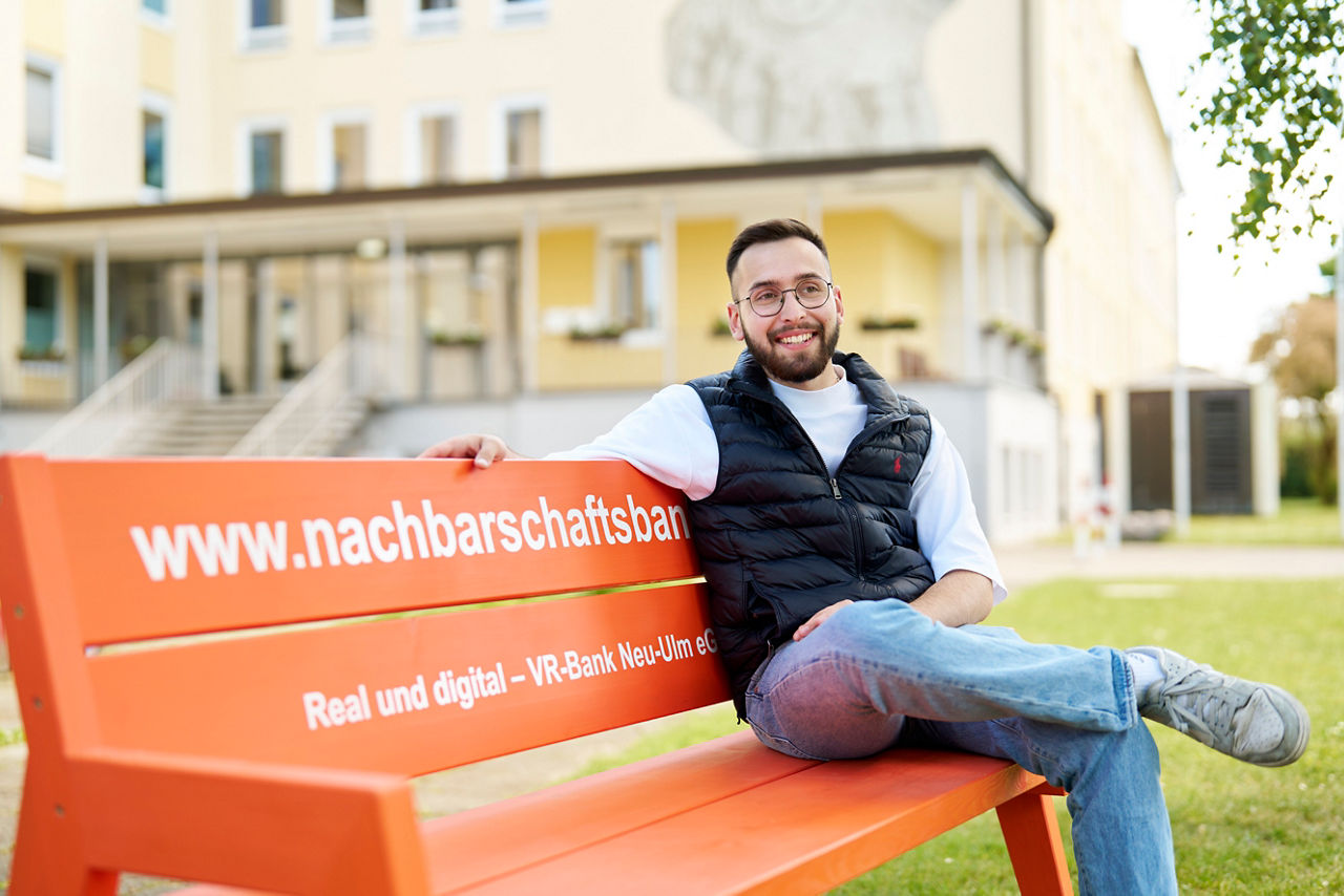 Max Müller sitzt auf einer orangen Nachbarschaftsbank und lacht in die Kamera