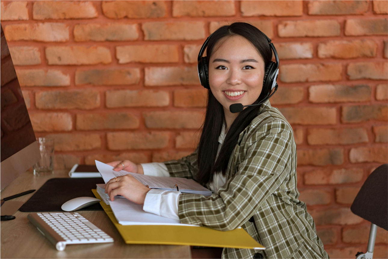 Eine Frau trägt ein Headset und sitzt vor einem PC. In der Hand hält sie einen gelben Ordner.