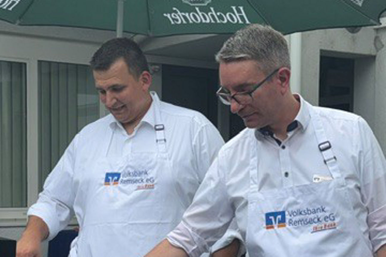 Zwei Männer in Schürzen der Volksbank Remseck eG grillen Würstchen unter einem grünen Hochdorfer Sonnenschirm.